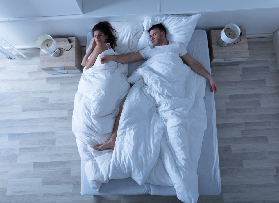 Overhead view of couple laying in bed, the man is asleep, his right arm is on the woman and she looks annoyed