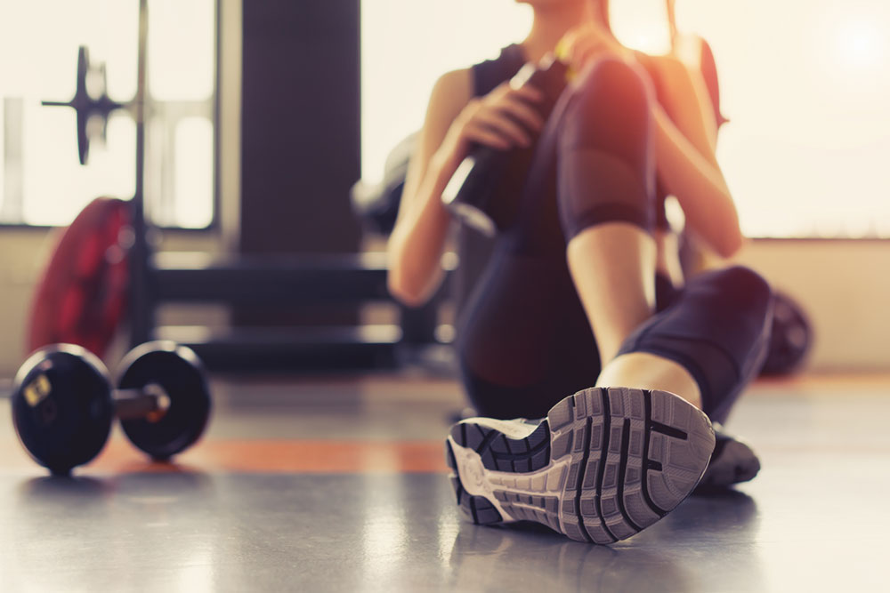 Female sitting on floor in the gym, one leg extended and blur image of dumbbell on floor next to her, water bottle in hand